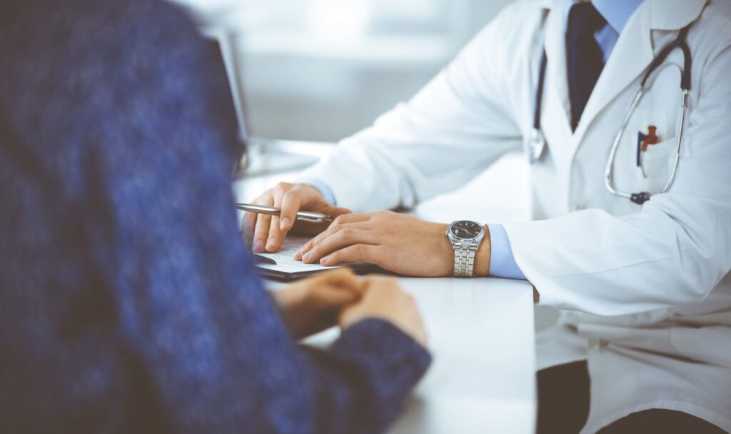 Unknown young woman patient discuss the results of her medical tests with a doctor, while sitting at the desk in a hospital office. Physician using clipboard for filling up medication history records. Perfect medical service in clinic.